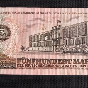 May include: A 500 Mark banknote from the German Democratic Republic. The banknote features a building with a red roof and a hammer and compass symbol in a circle.