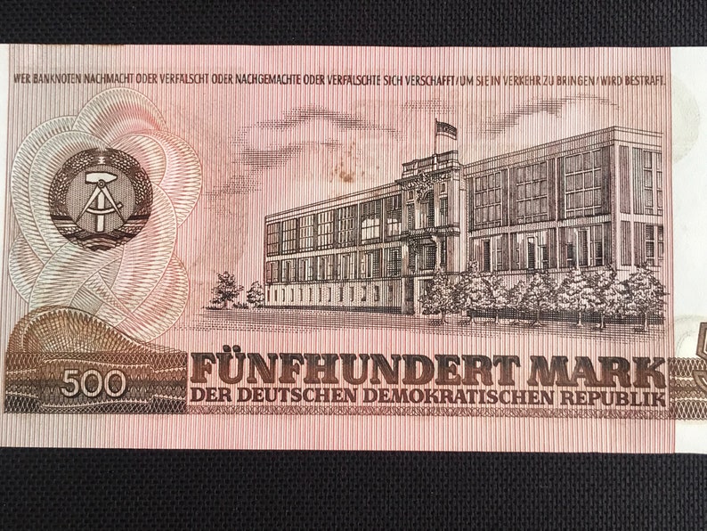May include: A 500 Mark banknote from the German Democratic Republic. The banknote features a building with a red roof and a hammer and compass symbol in a circle.