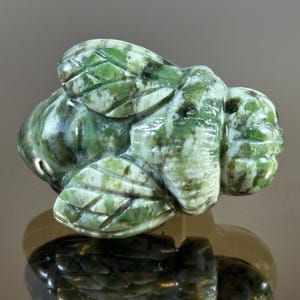 May include: A carved jade bee figurine. The bee is green and white with detailed wings and body segments. The figurine is displayed on a reflective surface, showcasing its intricate design and craftsmanship.