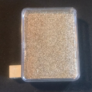 Könnte beinhalten: Ein durchsichtiger Plastikbehälter gefüllt mit einem hellbraunen, feinkörnigen Material. Der Behälter steht auf einer dunklen Oberfläche.