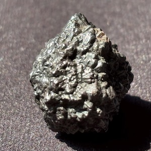 May include: A dark, textured, irregularly shaped rock with a rough surface. The rock is a dark gray color and appears to be metallic. The image is a close-up shot, highlighting the rock's unique texture and form.