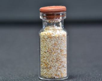 Könnte beinhalten: Eine kleine Glasflasche mit braunem Deckel, gefüllt mit weißem Sand.