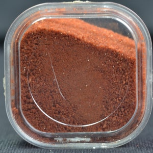 May include: A small clear plastic container filled with a reddish-brown powder.