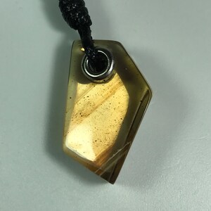 Puede incluir: Un colgante dorado con un collar de cordón negro. El colgante tiene una forma geométrica con un anillo de metal plateado en la parte superior.