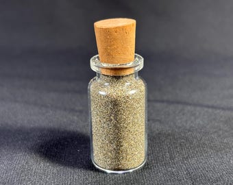 Puede incluir: Una pequeña botella de vidrio con un tapón de corcho llena de brillantina fina de color marrón claro.