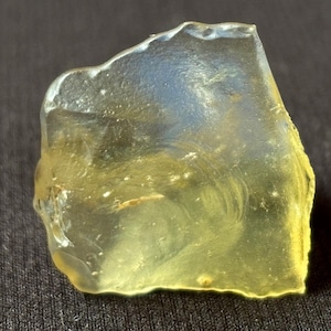 Könnte beinhalten: Ein durchscheinendes, unregelmäßiges Stück gelbes Glas mit einer glatten, polierten Oberfläche. Das Material weist interne Wirbel und lichtreflektierende Eigenschaften auf und zeigt einen blassblauen Schimmer in Bereichen, in denen Licht durch die Struktur fällt.