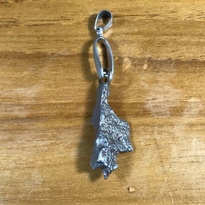 Puede incluir: Un colgante de plata con una superficie rugosa y texturizada en forma de roca irregular. El colgante tiene un bucle de plata en la parte superior para sujetarlo a una cadena.