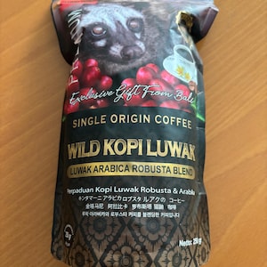 Könnte beinhalten: Ein dunkler Kaffeepackung mit einer Zibetkatzen-Illustration, roten Kaffeekirschen und dem Text "Exclusive Gift From Bali" und "Wild Kopi Luwak". Auf der Packung steht auch "Single Origin Coffee" und "Luwak Arabica Robusta Blend".