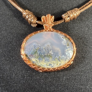 Könnte beinhalten: Ein Anhänger aus Kupferdraht, der einen hellgrauen Achat-Stein mit grünen und braunen Einschlüssen umwickelt. Der Anhänger ist an einer braunen Schnur befestigt.