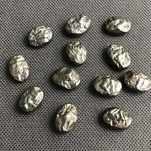 May include: A collection of 13 small, oval-shaped, silver-colored metal pieces with a textured surface. The pieces are arranged in a random pattern on a gray fabric background.