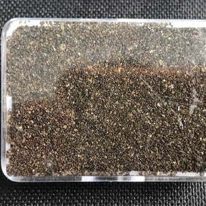 Peut inclure: Un récipient en plastique transparent rempli de petites particules de sable brun foncé et gris.