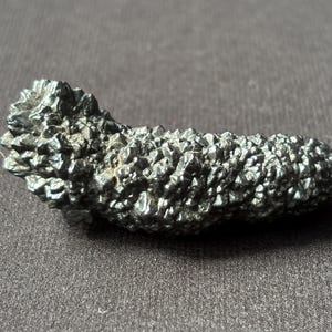 Pode incluir: Um espécime mineral cinza escuro, texturizado, com uma superfície áspera e cristalina. A forma alongada e o brilho metálico sugerem pirita ou um mineral semelhante. O espécime é exibido em um fundo cinza escuro.