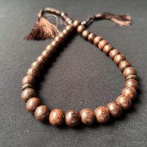 May include: A string of prayer beads made from dark brown wooden beads. The beads are round and smooth, with a natural wood grain pattern. A tassel of brown threads hangs from each end. The beads are arranged in a loop.