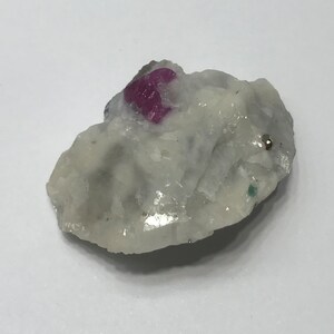 Puede incluir: Una pequeña roca cristalina blanca con un cristal de rubí rosa brillante incrustado en ella. Hay una pequeña mancha metálica dorada en la roca.