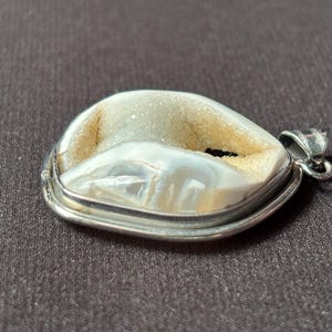 May include: A pendant featuring a polished, irregular-shaped stone with a smooth, creamy white outer edge and a textured, sparkling beige druzy center. The stone is encased in a simple, polished silver-toned metal setting with a small bail at the top.