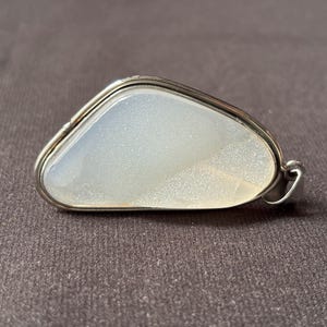 Può includere: Un ciondolo caratterizzato da una pietra bianca lattiginosa, traslucida e con un sottile riflesso iridescente, incastonata in una cornice di metallo color argento lucido. La pietra ha una forma triangolare irregolare e arrotondata.