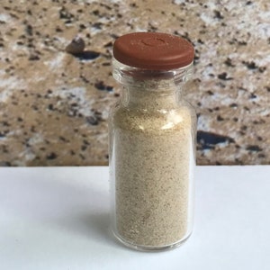 May include: A small, clear glass vial filled with a light tan granular substance. The vial has a brown cap and is set against a neutral background. The contents appear to be fine particles.