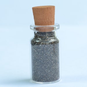 Puede incluir: Una pequeña botella de vidrio con un tapón de corcho llena de semillas de amapola negras.