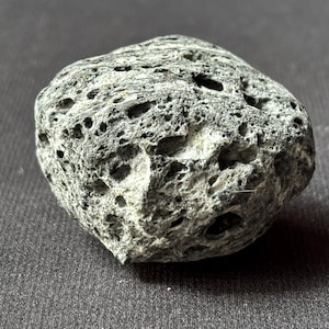 Puede incluir: Una roca volcánica gris porosa con una superficie rugosa y texturizada. La roca tiene numerosos agujeros pequeños y una forma ligeramente redondeada. El color es una mezcla de gris y blanco, con áreas más oscuras en los agujeros.