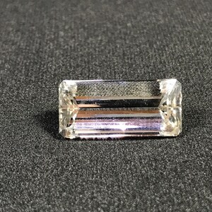 May include: A rectangular, clear gemstone with multiple facets, reflecting light. The gem is set against a dark gray background, highlighting its clarity and cut. The gemstone appears to be a high-quality, polished crystal.