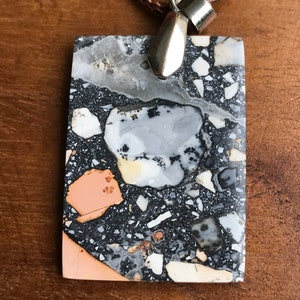 May include: A rectangular pendant made of terrazzo with a silver bail. The pendant has a black, white, and orange color scheme with a variety of small, round, and irregular shapes.