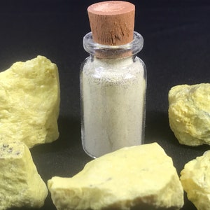 Può includere: Un piccolo flacone di vetro con un tappo di sughero riempito di polvere bianca. Il flacone è circondato da cinque pezzi di roccia gialla.