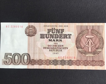 Op de afbeelding: Een bankbiljet van 500 Mark uit de Duitse Democratische Republiek, gedateerd 1985. Het biljet heeft een rood en bruin ontwerp met de tekst "Fünf Hundert Mark" en de woorden "Der Deutschen Demokratischen Republik" in het midden. Het biljet heeft ook een kompas- en hamersymbool.