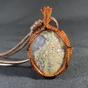 Puede incluir: Un colgante envuelto en alambre de cobre con una piedra de ágata musgosa. El colgante está unido a un cordón marrón.