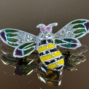 Puede incluir: Broche de abeja detallado con alas plateadas incrustadas con pequeñas piedras brillantes. Las alas tienen detalles de esmalte verde y morado. El cuerpo de la abeja es de esmalte amarillo y negro. El broche tiene un cierre de alfiler.