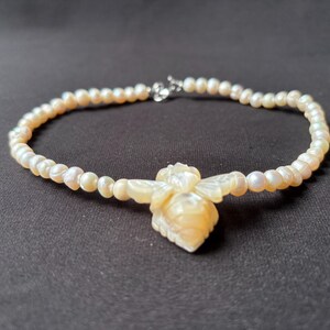 Könnte beinhalten: Zarte Halskette mit einer Kette aus kleinen, cremeweißen Perlen. Der Anhänger ist eine geschnitzte, cremefarbene Biene. Der Verschluss ist silberfarben. Der Schmuck ist vor einem dunklen Hintergrund platziert.