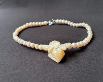 May include: A necklace made of small, creamy-white pearls, with a carved, off-white bee pendant. The clasp is silver. The jewellery is displayed against a dark background. The pearls are approximately 0.3 cm in diameter.