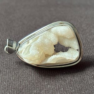 Könnte beinhalten: Ein tropfenförmiger Anhänger mit einem texturierten, kristallinen weißen Druzy-Stein, der in einen polierten, silberfarbenen Metallrahmen gefasst ist. Der Anhänger verfügt über eine kleine Metallöse an der Oberseite zur Befestigung an einer Kette.
