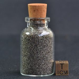 Può includere: Una piccola bottiglia di vetro trasparente riempita con materiale scuro e granulare, sigillata con un tappo di sughero. Un piccolo cubo di bronzo con il testo "T" e "1CM" è accanto alla bottiglia. Sfondo scuro.