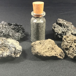 May include: A small glass bottle with a cork stopper filled with black volcanic sand. Five pieces of black and gray volcanic rock are arranged around the bottle on a dark gray surface.
