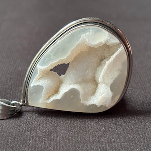 Puede incluir: Un colgante en forma de lágrima que presenta una piedra de ágata blanca translúcida con una cavidad de geoda central llena de formaciones de drusa blanca cristalina y brillante, todo engastado en un marco de metal plateado pulido con una reasa.