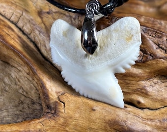 May include: A shark tooth pendant necklace. The pendant is a large, off-white shark tooth with a silver-colored metal bail. The necklace has a black cord. The background is a piece of wood.