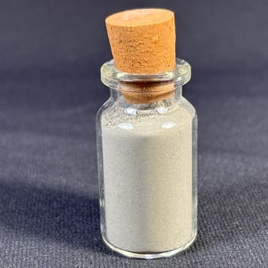 May include: A small glass bottle with a cork stopper, filled with a light gray powder.