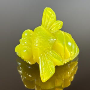 May include: A detailed, yellow-green carved sculpture of a stylized insect, possibly a bee or a beetle. The sculpture has intricate details, including wings and segmented body parts. The object is highly reflective, with a smooth, polished surface.