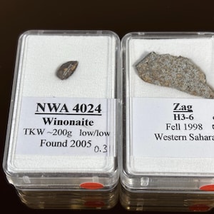 Pode incluir: Dois meteoritos em caixas de exibição transparentes. A caixa esquerda contém um pequeno meteorito escuro, em forma oval, rotulado como "NWA 4024 Winonaite". A caixa direita contém um meteorito maior, de forma irregular, rotulado como "Zag H3-6 Fell 1998 Western Sahara."