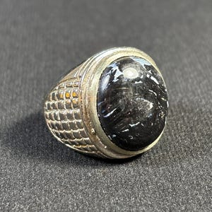Puede incluir: Un anillo de plata con una piedra preciosa negra. El anillo tiene una banda texturizada y un engaste redondo.