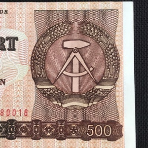 May include: A brown and white banknote with a brown and white emblem of a compass and hammer surrounded by wheat stalks. The text "500" is printed in brown on a black band at the bottom of the banknote.