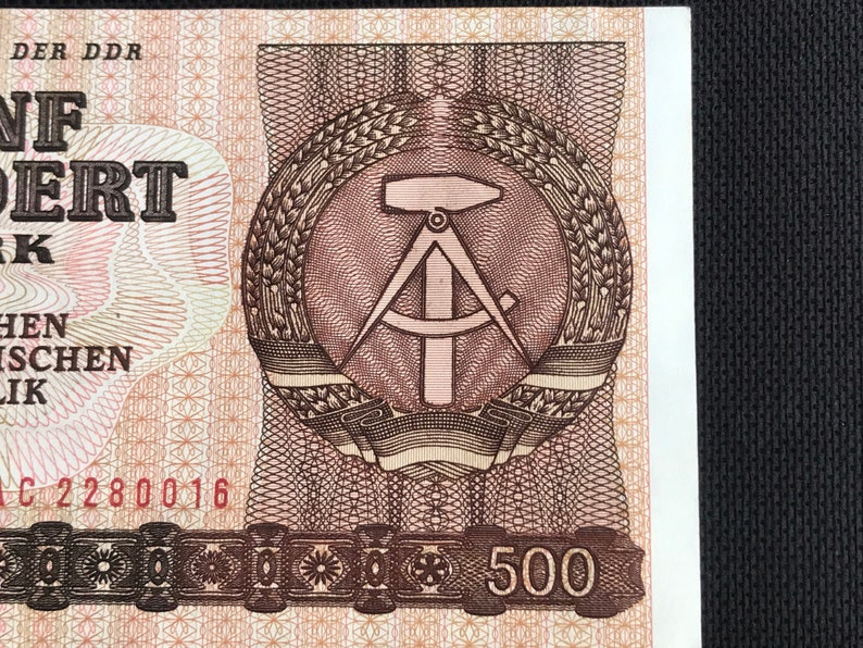May include: A brown and white banknote with a brown and white emblem of a compass and hammer surrounded by wheat stalks. The text "500" is printed in brown on a black band at the bottom of the banknote.