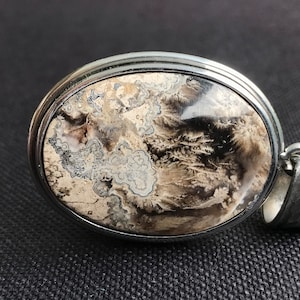 Könnte beinhalten: Ovaler Anhänger mit silberfarbenem Metallrahmen. Der Anhänger zeigt einen Naturstein mit einem Muster aus beige, grauen und schwarzen Wirbeln und federartigen Texturen. Die Öse ist ebenfalls silberfarben.