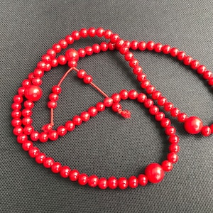 May include: A red beaded necklace with a tassel. The beads are round and uniform in size. The necklace is made of coral.