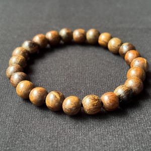Może przedstawiać: Bransoletka z okrągłych drewnianych koralików w różnych odcieniach brązu. Koraliki mają widoczne usłojenie drewna. Elastyczna bransoletka jest pokazana na czarnym tle. Koraliki mają średnicę około 1 cm.