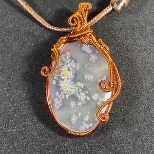 Puede incluir: Un colgante envuelto en alambre de cobre con una piedra de ágata blanca con un patrón dendrítico. La piedra tiene forma ovalada y tiene un fondo gris claro con dendritas marrones y blancas.