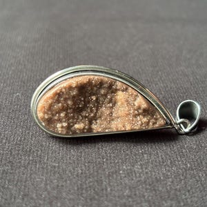 May include: A teardrop-shaped pendant featuring a textured, light brown druzy stone set within a polished silver-toned metal frame. The pendant includes a small, integrated silver-toned metal bail at the pointed end for attachment to a chain.