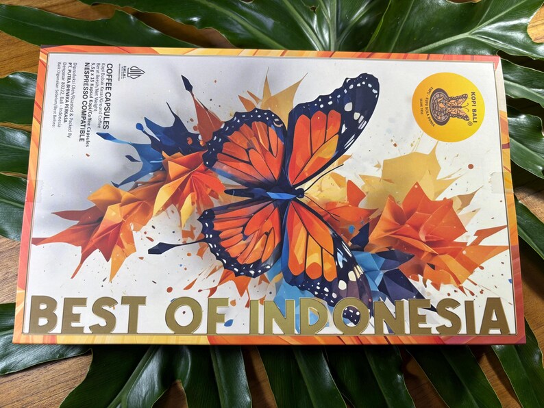 Puede incluir: Una caja de c&aacute;psulas de caf&eacute; con un dise&ntilde;o colorido que presenta una mariposa y el texto "BEST OF INDONESIA". La caja tiene un sello dorado que dice "KOPI BALI". El fondo tiene formas geom&eacute;tricas naranjas, azules y amarillas.