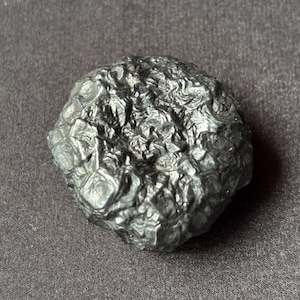 Puede incluir: Una piedra de color gris oscuro, de forma irregular, con una superficie texturizada. La piedra tiene un aspecto rugoso y con bultos y parece metálica. El objeto está sobre un fondo gris oscuro.