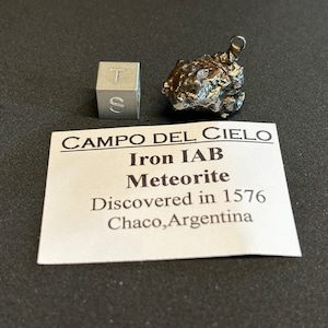 May include: A small, dark grey meteorite with a silver loop attached. The meteorite is labelled "Campo del Cielo Iron IAB Meteorite Discovered in 1576 Chaco, Argentina."